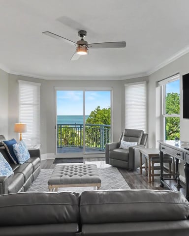 Bright living room with gray leather seating, ocean and tree views through glass doors, wall-mounted TV, and ceiling fan.