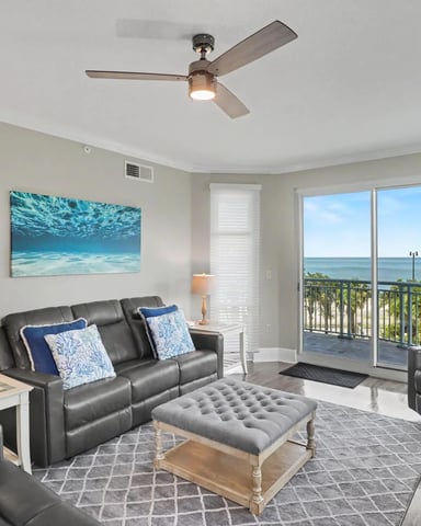 Beachfront living room with gray leather seating, ocean view through glass doors, and ceiling fan