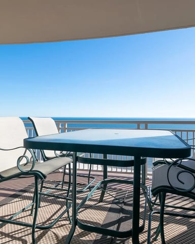 Oceanfront balcony with metal dining table, cushioned chairs, and clear blue sky view