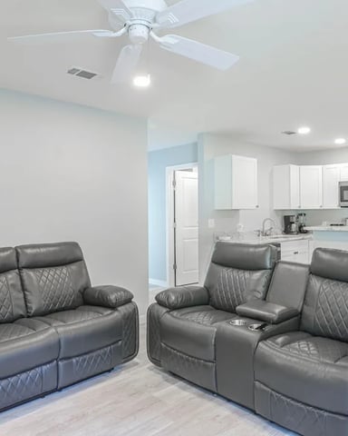 Open-plan living room with two gray leather recliners facing a kitchen with white cabinets and stainless steel appliances