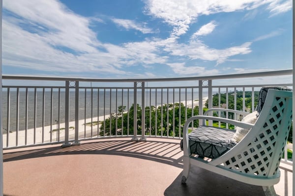Curved balcony with white railing overlooking sandy beach, trees, and water under blue sky with clouds