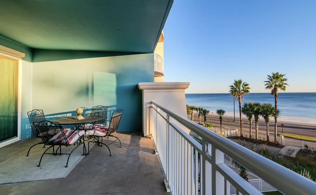 Oceanfront balcony with wrought-iron dining set, teal walls, and views of palm trees, a coastal road, and the sea