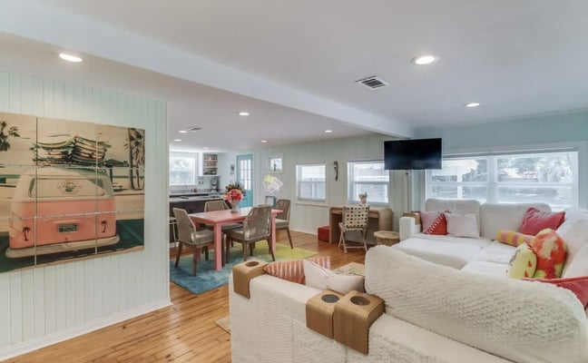 Open-plan coastal living and dining room with white sectional, pink VW bus wall art, wood floors, and pastel beach decor