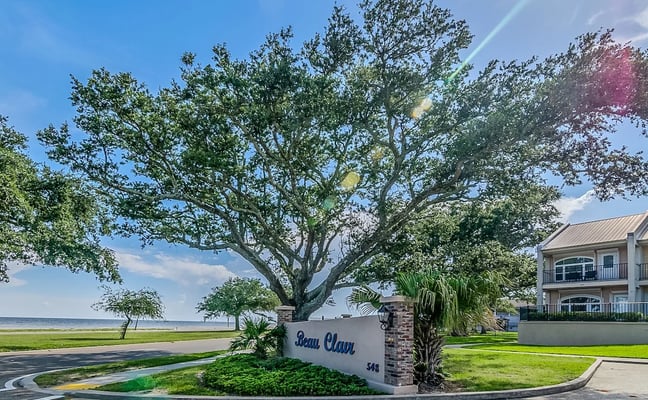 Waterfront property entrance with large oak tree, white sign reading Beau Clair, and two-story residential building overlooking bay